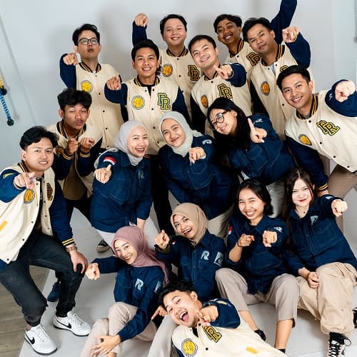 Tim RRbrother berfoto bersama mengenakan jaket varsity krem dan seragam biru sambil mengangkat tangan ke arah kamera