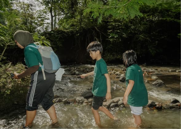 Rombongan pelajar berjalan beriringan menyusuri jalur setapak di tepi sungai berbatu di dalam hutan