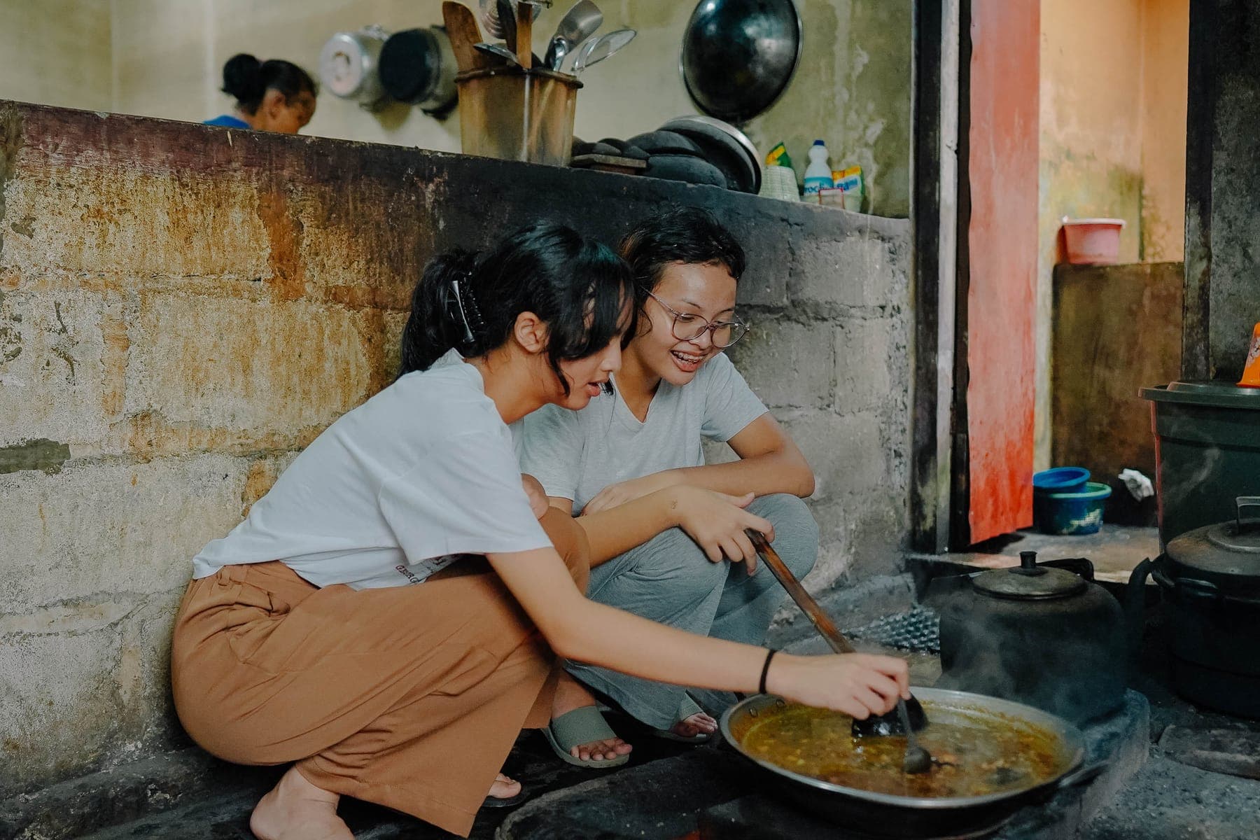 Seorang siswa berjongkok di depan tungku kayu sambil memasukkan kayu bakar, dibimbing ibu tuan rumah