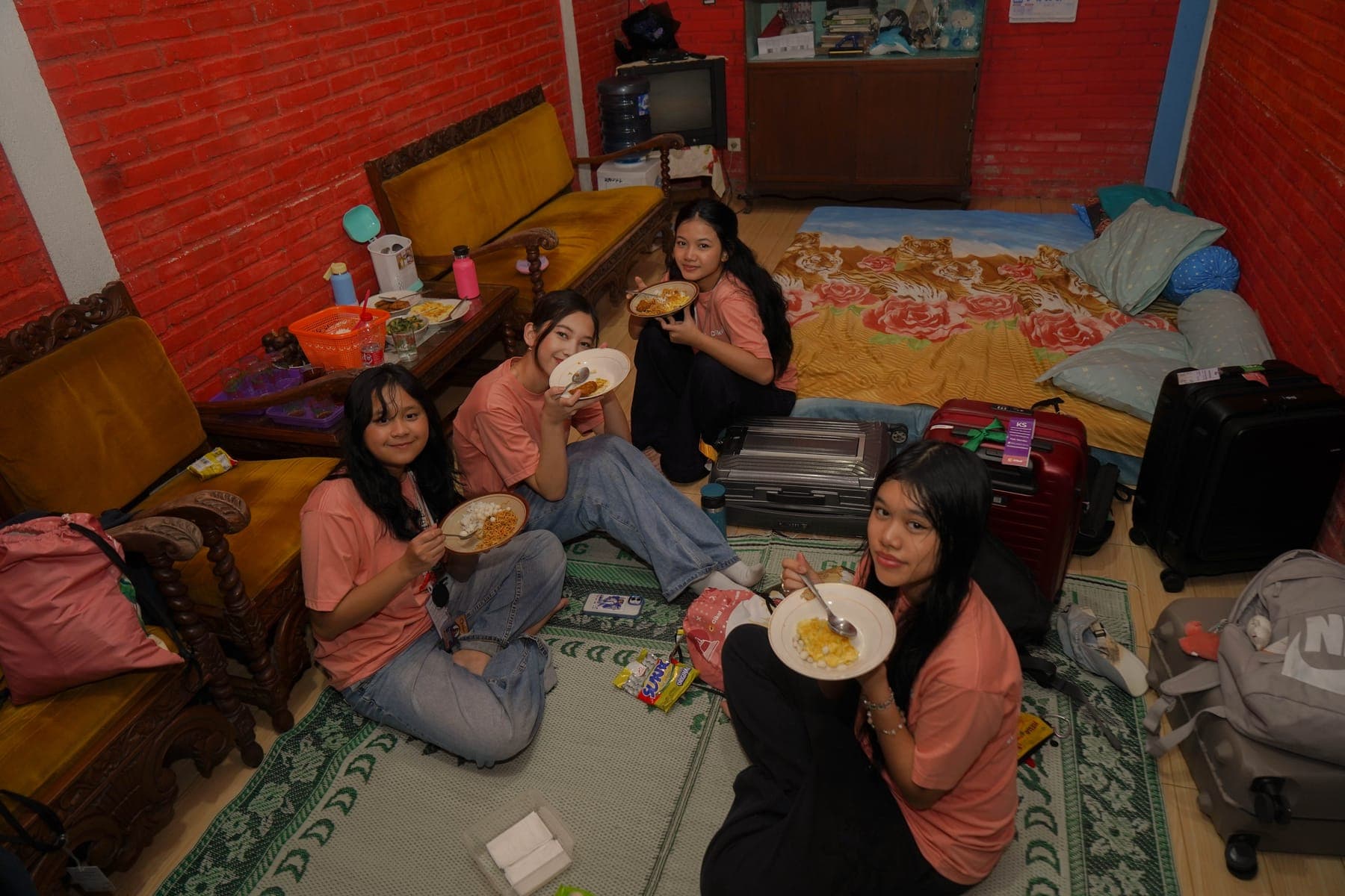Beberapa siswa makan bersama beralaskan tikar di ruang tengah rumah warga, memakai piring dan gelas milik tuan rumah